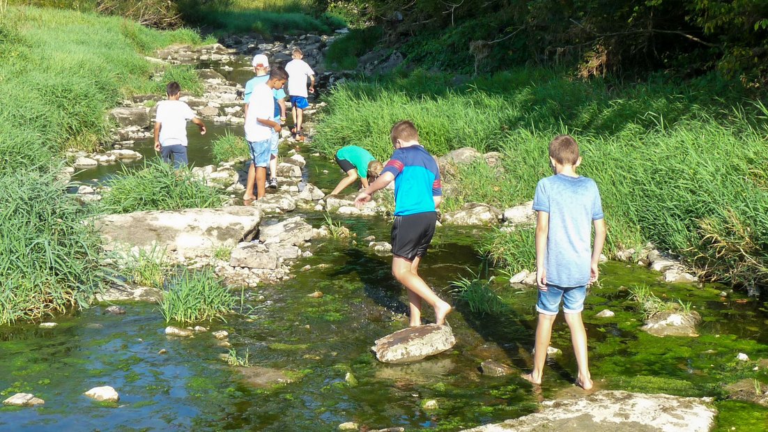 Mlađi učenici istražuju plitko korito reke sa velikim kamenjem tokom dana pešačenja.